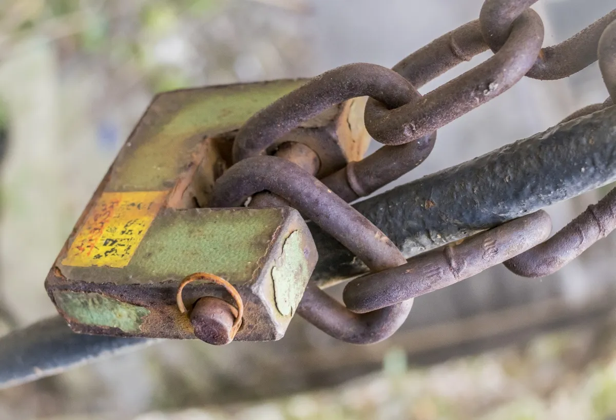 Rusty chain looped through a weathered green padlock on a metal bar